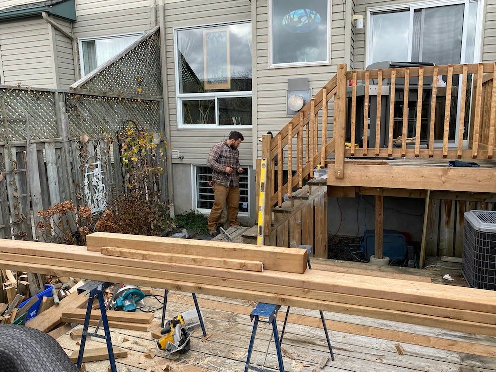 Raised deck with a railing under construction behind a house, lumber and saw horses in the foreground and a man working below