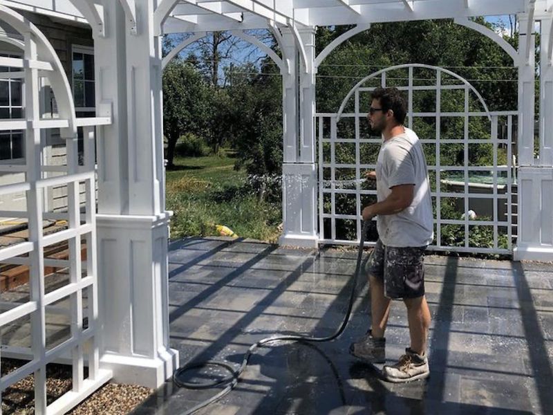 White pergola with arched lattice panels over a stone patio, a man rinsing the floor with a hose