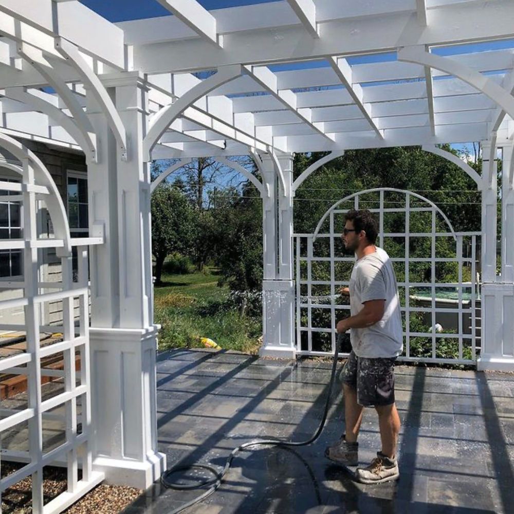 Inside a white pergola with a slatted roof and arched lattice panels, a man holding a hose on the stone floor