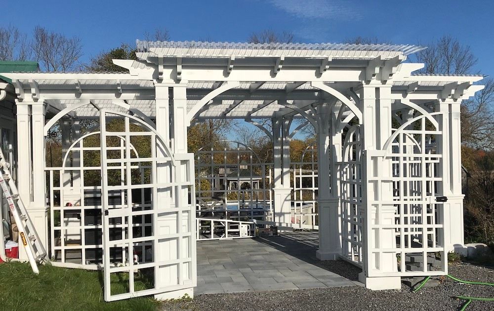 White pergola with a slatted roof, arched lattice walls and open lattice doors on a paved floor, a ladder against the side