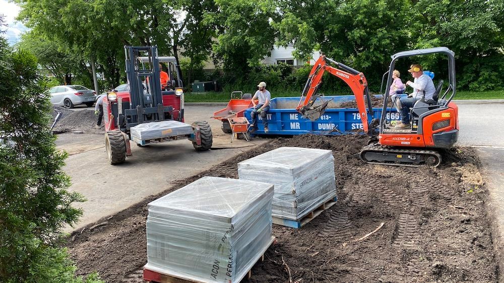 Orange mini excavator, a forklift and a dump bin beside two wrapped pallets of paving stones on a cleared strip of ground