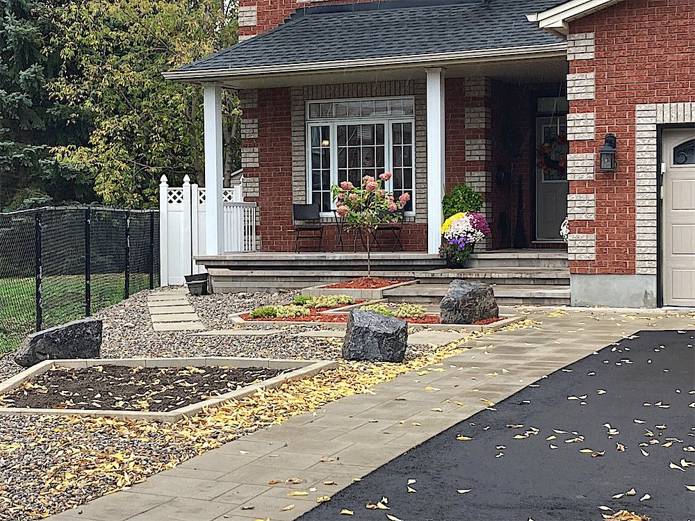 Paver front walkway beside an asphalt driveway, with large boulders and gravel beds in front of a brick house