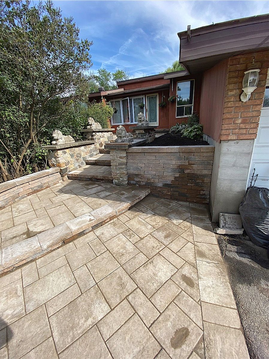 Beige paver walkway in a herringbone layout rising to stone steps and a stone retaining wall beside a house