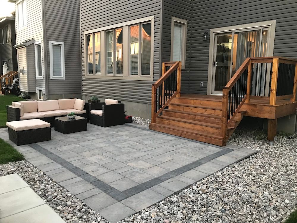 Grey paver patio with a dark border and outdoor sofas below a cedar deck with black railings and wide steps, gravel along the edge