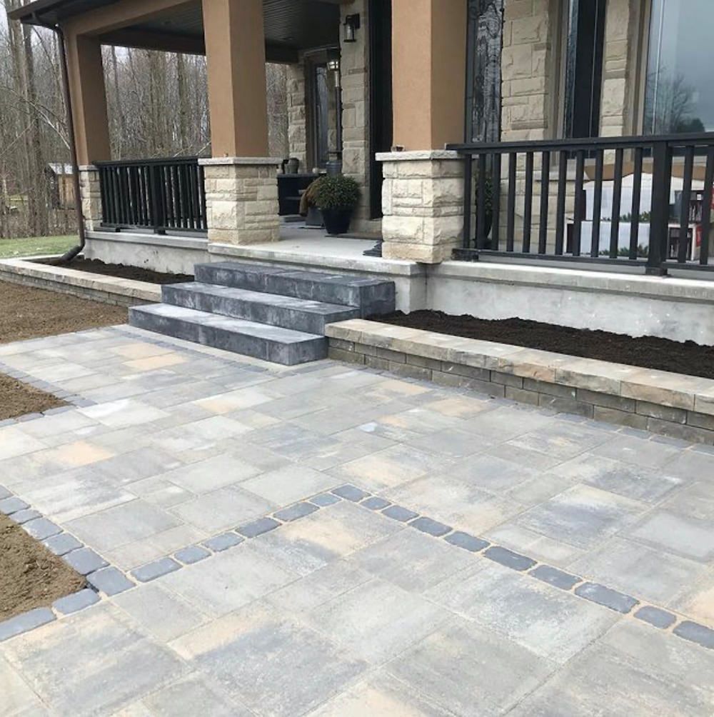 Multi-tone paver patio with a cobble inlay line in front of wide stone steps and a covered porch with black railings