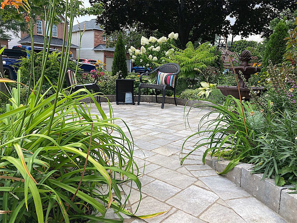 Light paver patio edged with a low stone border and full garden beds of daylilies, hydrangeas and ferns, a bistro chair at the far side