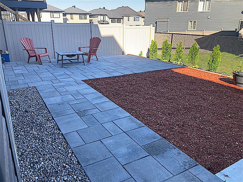 Blue-grey paver patio with a strip of grey gravel and a bed of red mulch, two red chairs and a fire bowl by a tall fence