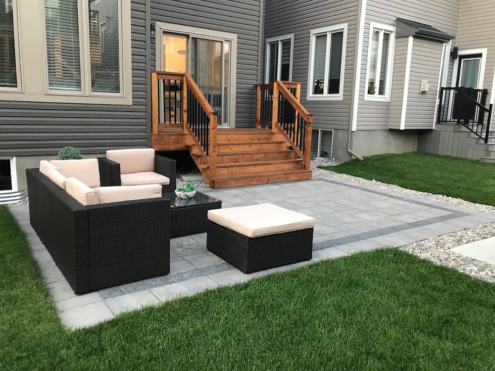 Black wicker lounge set with cream cushions on a grey paver patio, a cedar deck with black railings behind, lawn in the foreground