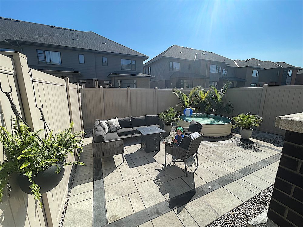 Light paver patio with a dark inlay line, an outdoor sofa and chairs, potted ferns and a round tub by a tall tan fence