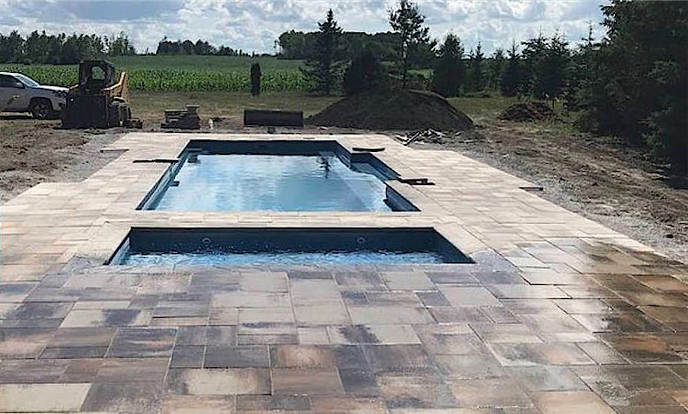 Rectangular in-ground pool with a wide paver deck around it, bare ground and trees behind