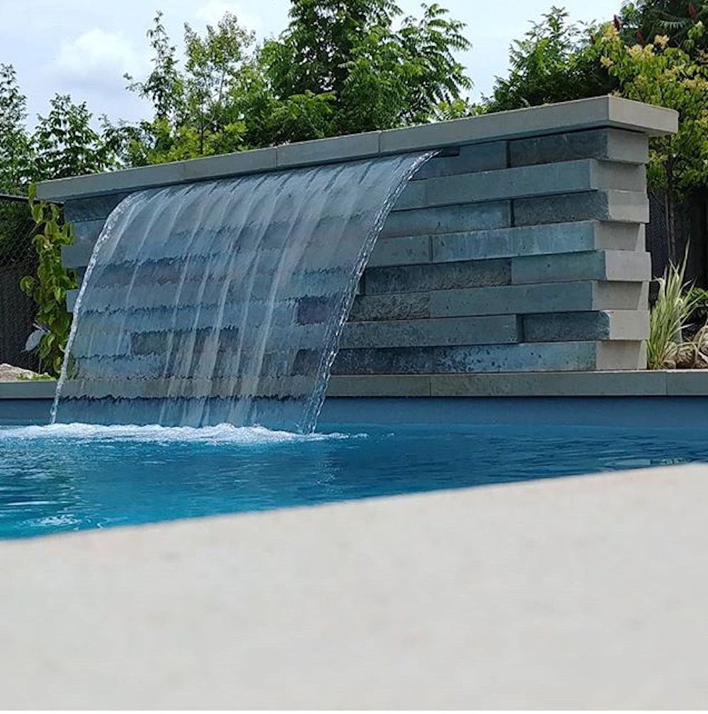 Stone-faced wall with water spilling over its top into a blue pool, a pale pool edge in the foreground