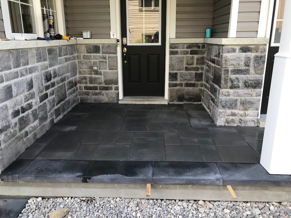 Dark paved porch floor between two stone-clad walls at a black front door, gravel along the front edge