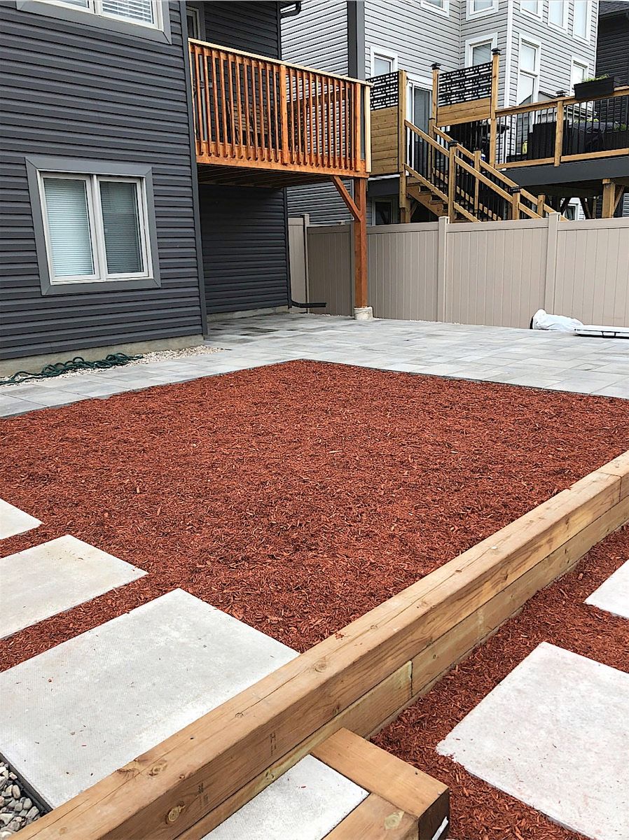 Large square stepping stones set in red mulch beside a paver area, with a timber edge in the foreground and a deck behind
