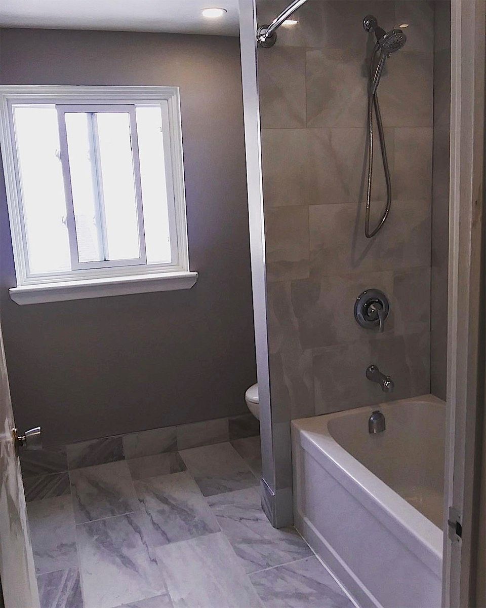Bathroom with a white tub, grey tiled shower wall, marble-look floor tile and a window