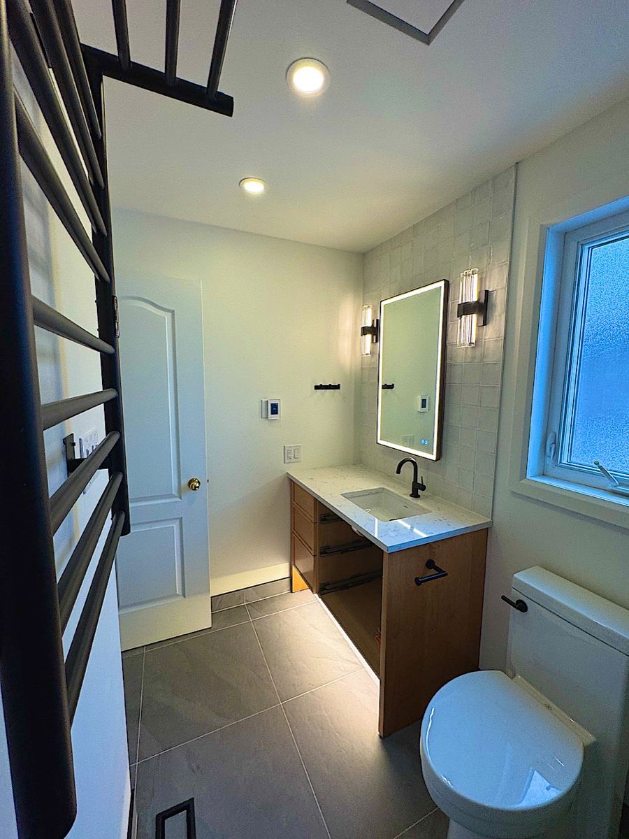 Bathroom with a wood vanity, a lit mirror, black fixtures, a black towel rack and a window beside the toilet