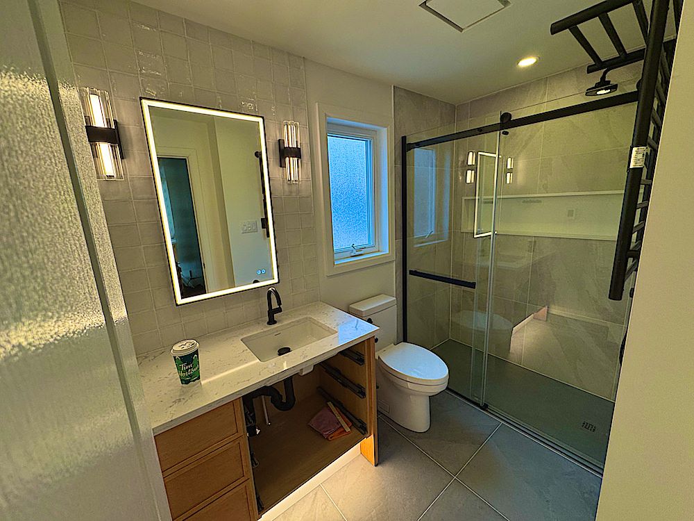 Bathroom with a wood vanity, a backlit mirror, wall sconces and a glass shower with a black frame
