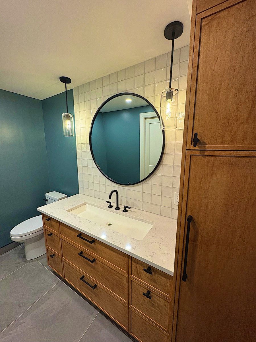 Bathroom with a wood vanity and drawers, a round black-framed mirror, pendant lights, square white wall tile and teal paint