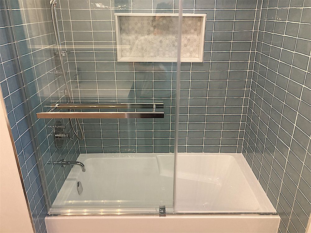 Bathtub with a glass door, blue-grey wall tile and a recessed tile shelf