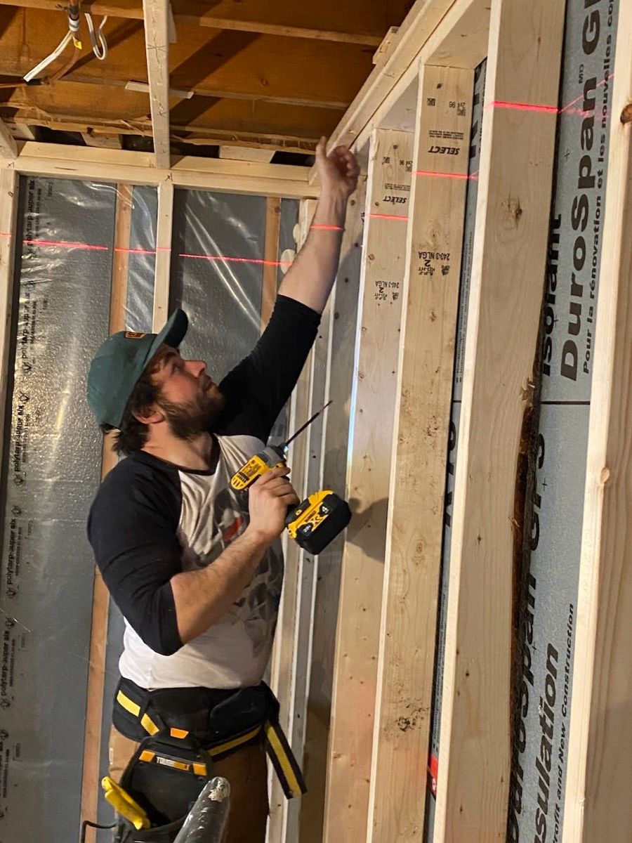Man in a ball cap drilling into new stud framing in a gutted room, laser level lines on the wall