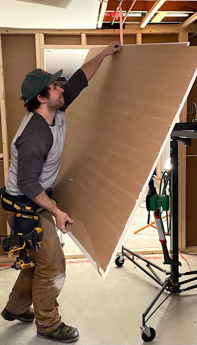 Man in a ball cap lifting a sheet of drywall on a panel lift against framing in a room being renovated